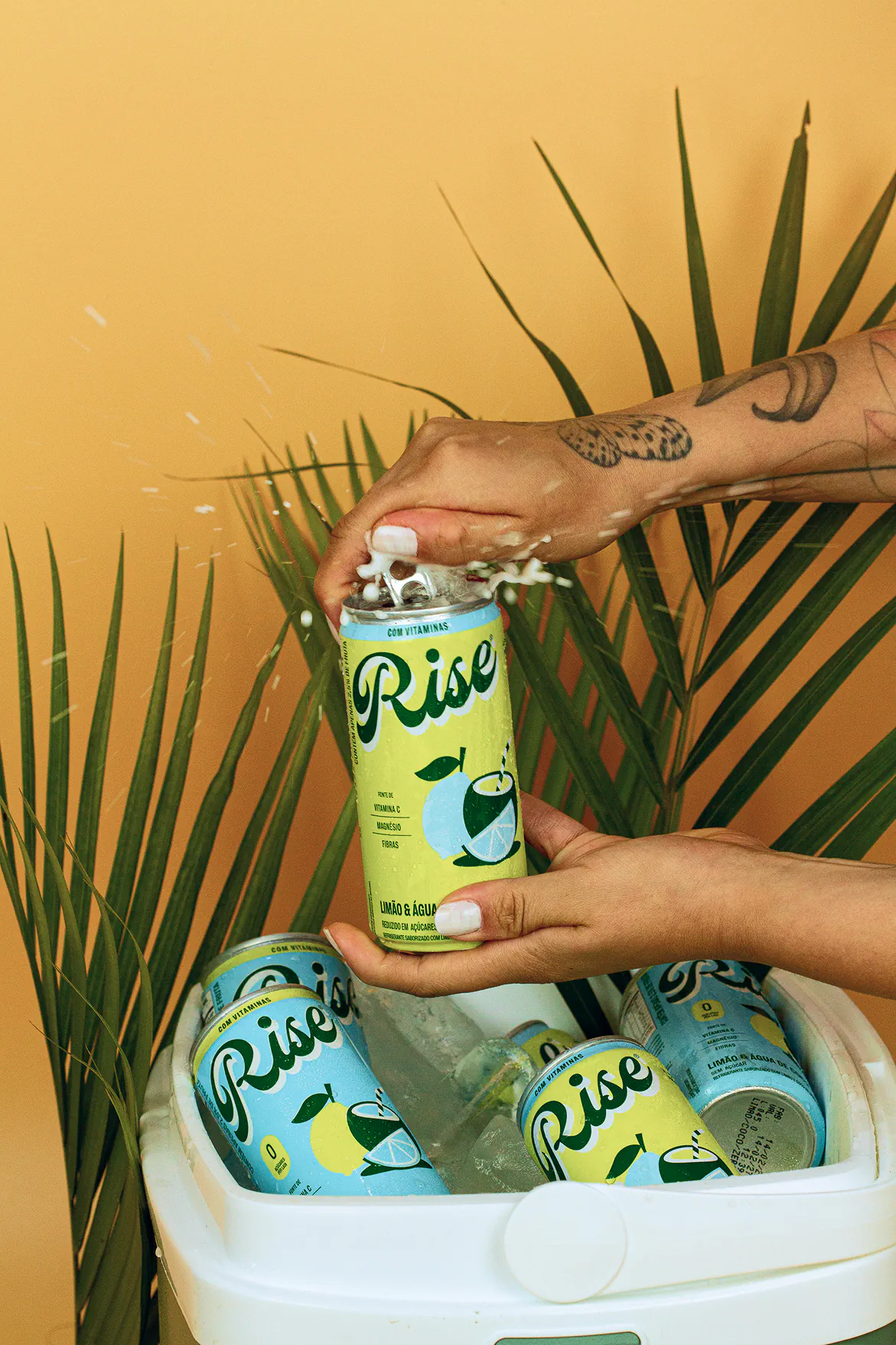 Mãos abrindo lata Rise Limão & Água de Coco com espuma, sobre cooler com gelo